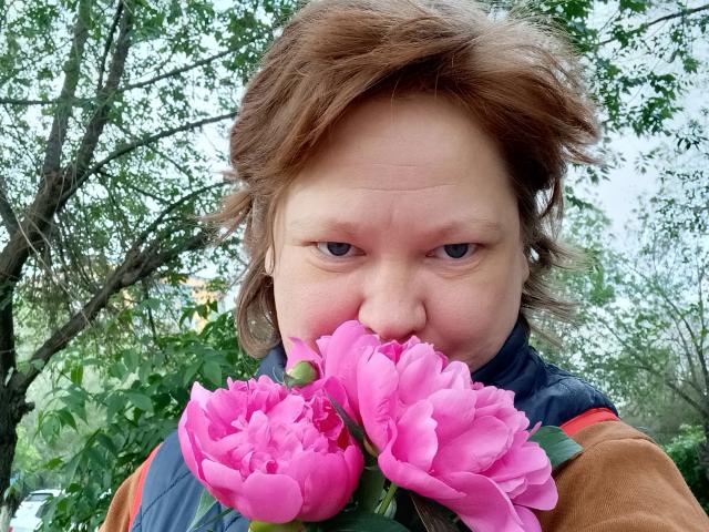 A close-up of DaysieFannyy with short, messy brown hair, holding pink peonies in front of her face. She has a slightly squinty, intense expression. Her blue and brown jacket is visible. The bright pink peonies contrast with her pale skin. The green leaves in the background are blurred. The photo has a natural, slightly overcast light. The focus is on DaysieFannyy's face and the peonies. The image has a casual, realistic style.
