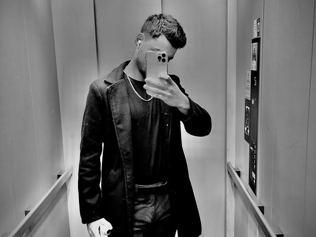Black and white photo of JamesFrenchie with short dark hair, wearing a dark jacket and shirt, taking a selfie in a narrow hallway, phone covering his face.