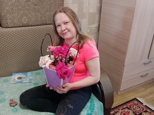 DaysieFannyy with light brown hair, pink shirt, and black pants, holding a purple vase with pink and white roses, sitting on a green-patterned table, smiling.