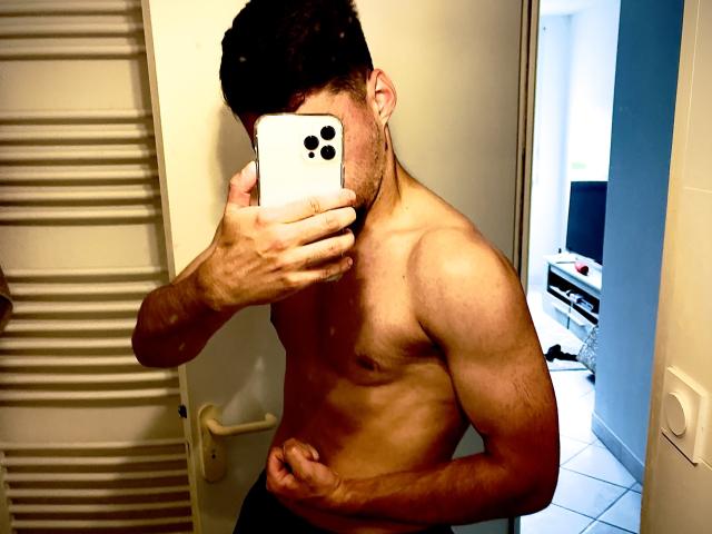 JamesFrenchie, shirtless with a muscular, hairy chest, holds a phone covering his face. His right hand is on his belly, showing off his toned abs and dark hair.