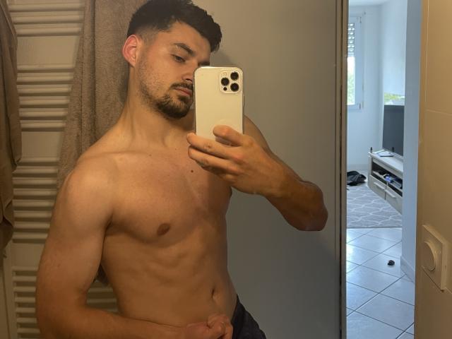 JamesFrenchie, shirtless, muscular, holding phone in mirror selfie, dark curly hair, beard, visible penis, standing in bathroom, towel on radiator, holding phone with white case.
