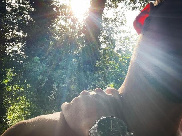 JamesFrenchie's sunlit profile with hand on neck, wearing a black wristwatch with a silver face, sunbeams through tree leaves, right side of face, red hair in background.
