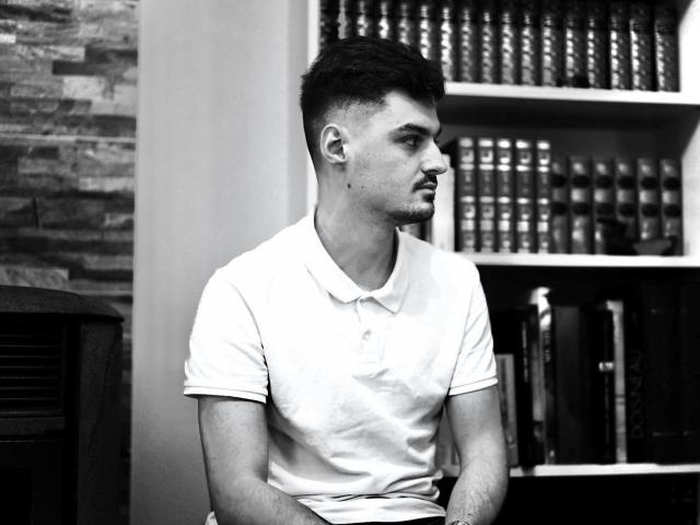 Black and white photo of JamesFrenchie, muscular, short dark hair, white polo shirt, looking to the right, bookshelf in background, small mole on left cheek, serious expression.
