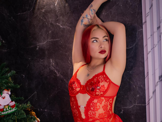 ViolettRoux with red lace lingerie, arms raised, showing tits, red hair, tattoos, and red lipstick, standing against dark marble wall. Christmas tree on left.