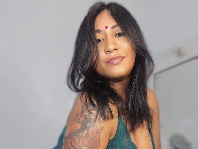 Nahirr with medium brown skin, black hair, and a red bindi on her forehead. She's smiling, wearing a green lace top, and has a colorful tattoo on her right arm.