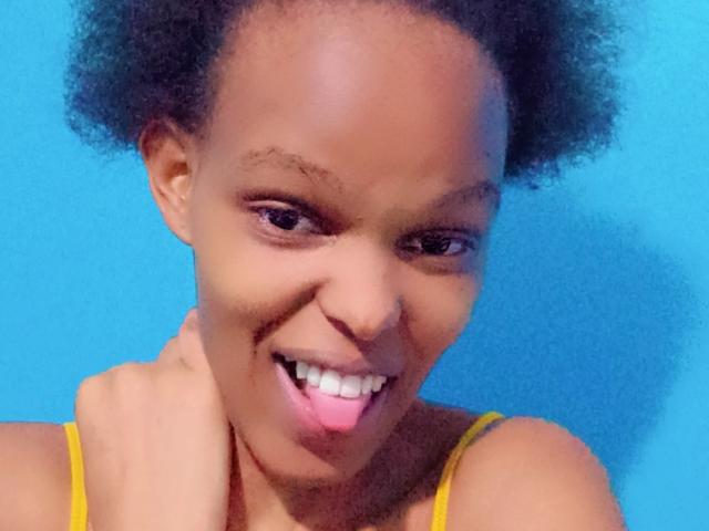 Pascalinee with dark curly hair, smiling with pink tongue out, yellow strap dress, hand on neck, blue background, brown skin, and small nose.