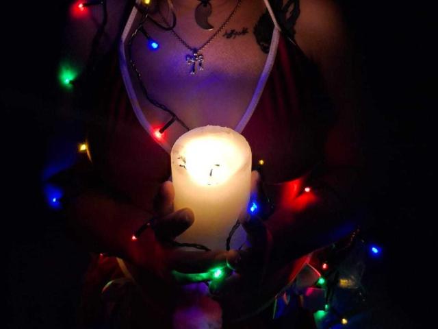 GraceRedMiller holding a lit candle with colorful Christmas lights wrapped around her chest, hands, and red bra, visible tits glowing softly in the candlelight.