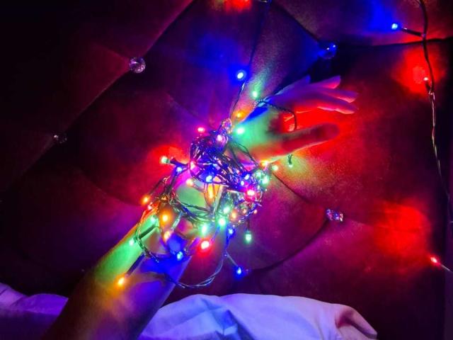 GraceRedMiller's hand holds colorful Christmas lights, glowing red, green, blue, and yellow, with purple and orange lights reflected on brown, tufted leather. Her other hand rests on white.