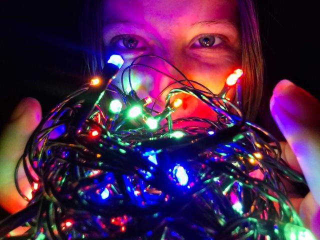 GraceRedMiller's face is lit by colorful Christmas lights, holding a tangled mess of blue, green, red, and yellow bulbs in front of her, eyes looking forward.