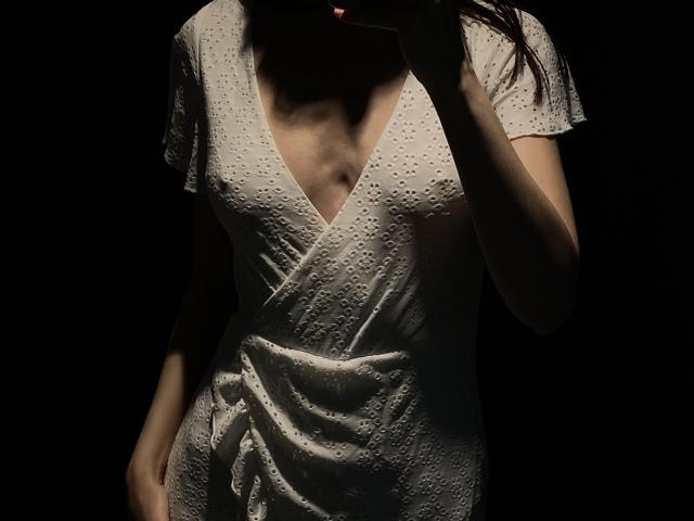 Valnes wears a white, deep V-neck, short-sleeved, textured dress, exposing her small tits and slight cleavage. Her right hand touches her dark, partially visible hair.