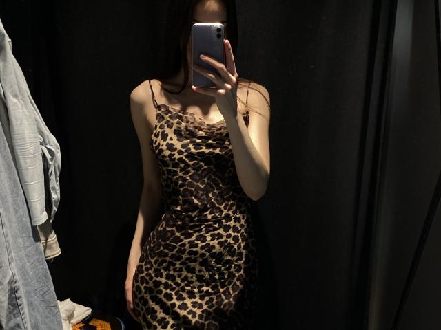 Valnes wearing a leopard-print dress, holding a phone in front of her face, showing her tits and ass, standing in a dimly lit room with dark curtains.