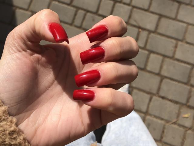 La mano de Valnes, con uñas rojas y brillantes en forma de almendra, exhibe sus pechos bien cuidados y brillantes. Lleva un suéter marrón claro y está de pie sobre una acera de ladrillo..