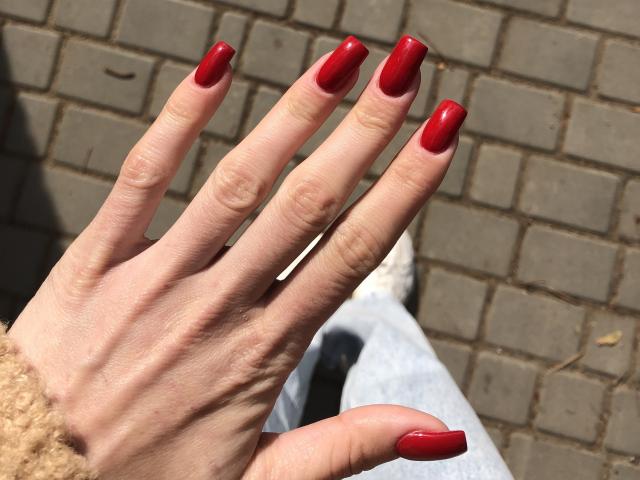 Valnes' hand with shiny red nails, fingers slightly spread, showing off her perfect, polished, glossy red boogers against a brick-patterned background.