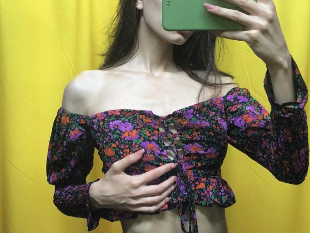 Valnes vestindo uma blusa floral com um ombro de fora e botões, segurando o celular na frente do rosto, mostrando seus pequenos seios e cintura fina, contra um fundo amarelo..