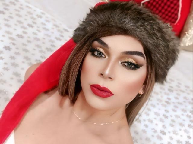 KarolElegance with red lipstick, green eyes, and a brown fur hat, looking up with a red Santa-like scarf. Her tits are barely visible, wearing a white top.