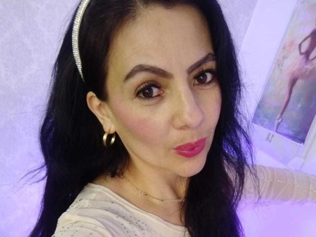 JakyKidman, avec ses longs cheveux noirs, portant un serre-tête scintillant, des boucles d'oreilles dorées, du rouge à lèvres rose et une chemise blanche, a un visage mignon et une expression légèrement boudeuse..