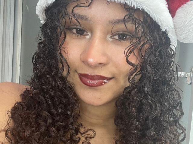 SofiaPassionne with curly brown hair, wearing a red Santa hat, has dark red lipstick and a silver lip piercing, smiling at the camera.