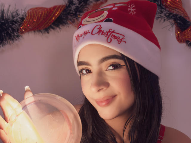 KonanStar with dark hair, wearing a red Santa hat, holding a glowing orb, smiling, wearing eyeliner, red lips, white nails, Christmas decor in background.