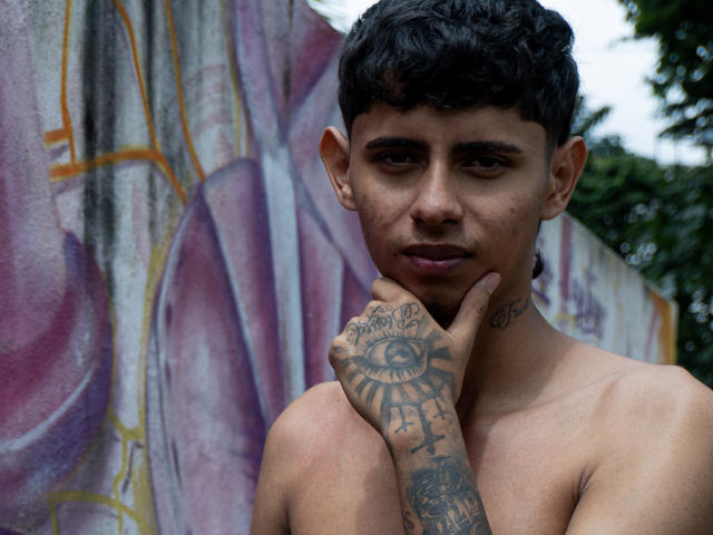 Shirtless JacobVillalobos with dark curly hair, tattoos on hand and neck, touches chin, serious expression, medium skin, standing against graffiti wall, green tree in background.