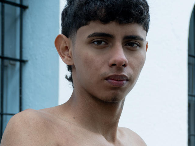 Shirtless JacobVillalobos with medium brown skin, short curly black hair, and a slight mustache, looking at the camera with a subtle, almost smirking expression.
