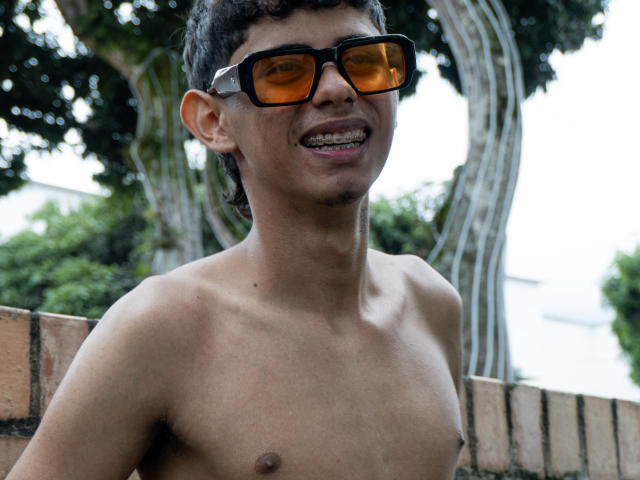 Shirtless JacobVillalobos with curly black hair, brown skin, small nipples, and orange sunglasses, smiling with metal braces, standing against a brick wall and tree.