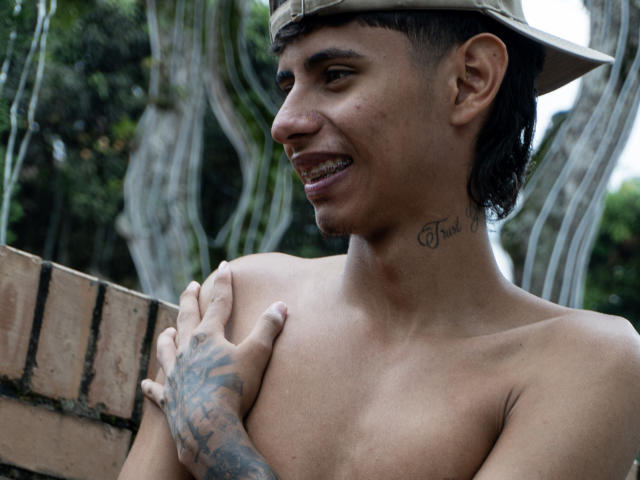 Shirtless JacobVillalobos with tattoos, wearing a hat, smiling, hand on chest, brown skin, short dark hair, tattoo on neck, forest background.