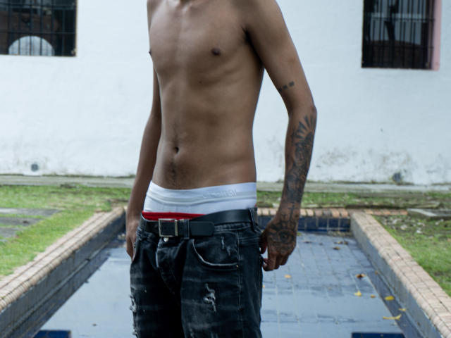 Shirtless JacobVillalobos with a slim, toned torso, dark arm tattoos, and black, worn jeans with a red and white waistband, standing by a small, rectangular pool.