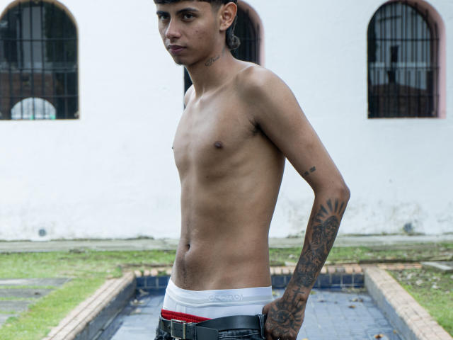 Slim, shirtless JacobVillalobos with tattoos, wearing white Calvin Klein underwear and black belt, standing with hands in pants, looking to the side.