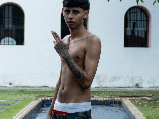 Shirtless JacobVillalobos with tattoos, pointing with right hand, wearing white Calvin Klein briefs, black belt, and red underwear, standing outside with a grassy and tiled area.