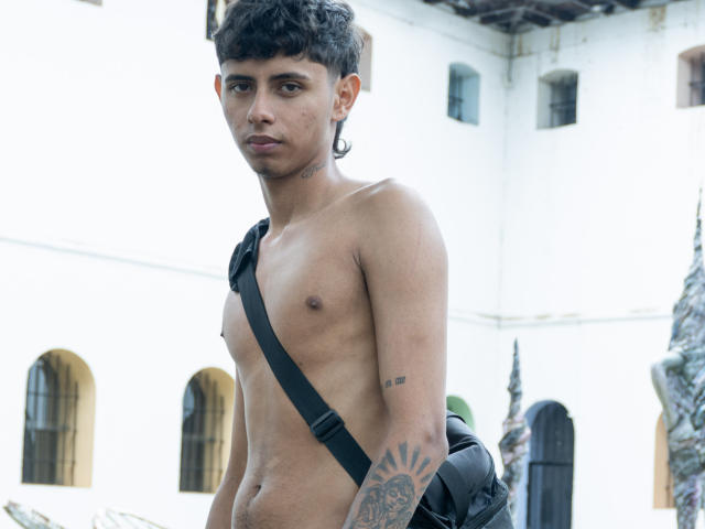 Shirtless JacobVillalobos with dark curly hair, small nipples, black strap, tattoos on arm, black earrings, serious expression, backpack, standing in daylight, white building in background.