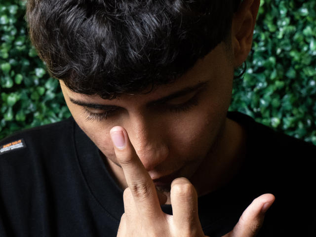 JacobVillalobos with dark curly hair, brown skin, and a black shirt, pressing his index finger to his nose, looking down, in a photo. His finger is near his nose.