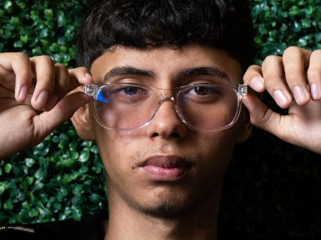 JacobVillalobos with dark curly hair, holding clear plastic glasses in front of his face, looking serious with a slight pout, against a green leafy background.