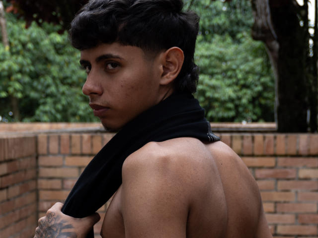 JacobVillalobos, shirtless, black scarf, tattoo on left hand, brown skin, short curly black hair, looking over shoulder, brick wall background, green trees, muscular back, serious expression.