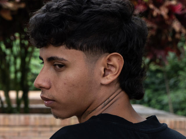 JacobVillalobos has curly black hair, a short undercut, a nose piercing, and a tattoo on his neck, wearing a black shirt, looking to the side with a serious expression.