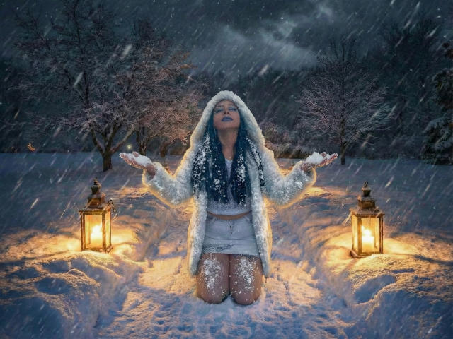 JessyLuxs kneels in snow, arms out, wearing a fluffy white hood, blue top, and white shorts, flanked by lanterns, snowflakes falling, dark night sky.