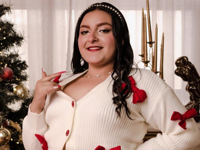VictoriaRiver with black hair, pearl headband, white cardigan with red bows, smiling, showing her tits, red nail polish, gold necklace, and Christmas tree in the background.