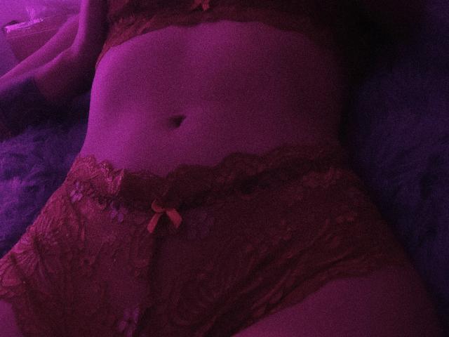 SofiaZen's pink-lit midsection showing her lace-covered pussy and lace-trimmed red panties with a small bow, lying on a fuzzy blue surface. Her navel is visible.