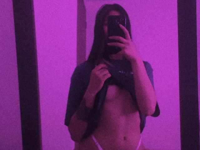 SofiaZen with long dark hair, lifting a shirt to reveal her small tits and white panties, taking a mirror selfie with purple lighting.