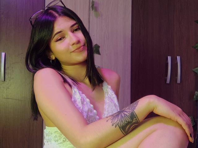 SofiaZen with black hair, wearing a white lace top, sitting with knees up, smiling, has a butterfly tattoo on her right arm, and is looking at the camera.