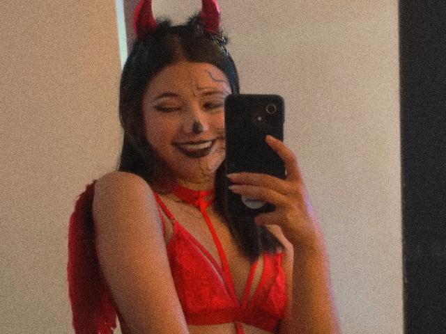 SofiaZen with red devil horns, red lace bra, and matching wings, smiling at a mirror selfie, holding a black phone, showing her tits and black lipstick.