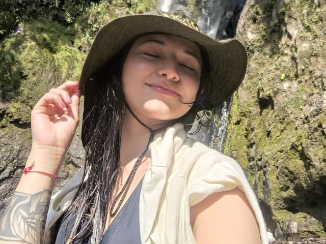 SofiaZen with wet black hair, wearing a straw hat and open shirt, smiling with eyes closed, showing tattoo on left arm, standing in front of a waterfall.