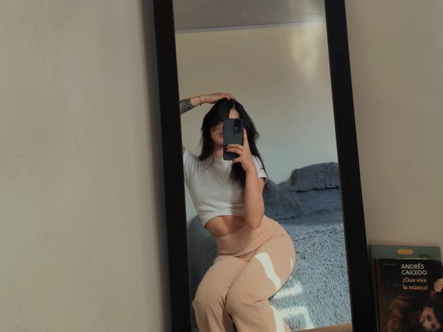 SofiaZen with long black hair, white crop top, and beige pants, sitting in front of a mirror, holding a phone, showing her curvy ass and small tits.