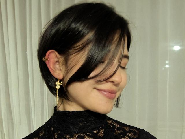 SofiaZen with short black hair, a gold earring, and a lace top, smiling with closed eyes. Her pink lips are slightly upturned, showing a gentle, cute expression.