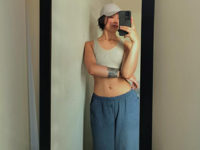 SofiaZen with short dark hair, wearing a white cap, gray sports bra, showing her tits, blue loose pants, holding a phone, tattoo on her left arm, standing in front of a mirror.