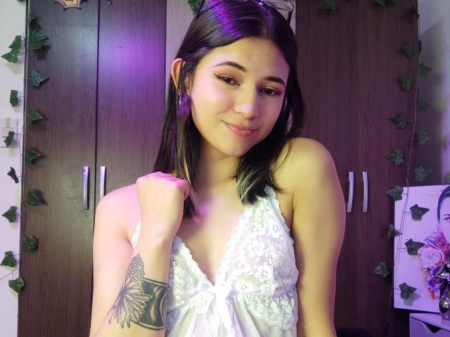 SofiaZen with dark straight hair, wearing a white lace dress, smiling, holding her hair, purple light on her, tattoo on her right arm, and eyeglasses on her head.