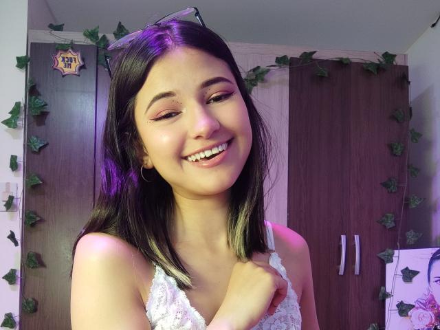 SofiaZen has medium brown skin, black and blonde ombre hair, and is smiling with her hand on her white lace top, showing her small tits and cute teeth.