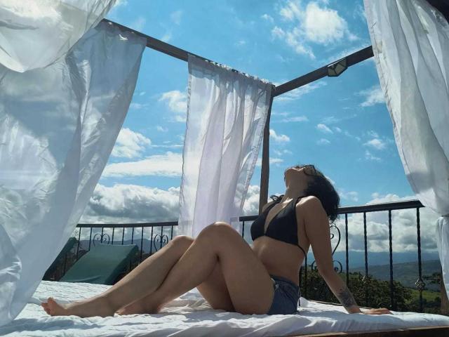 SofiaZen sits on a bed with white curtains, wearing a black bikini top and blue shorts, her back arched, eyes closed, sunlight on her tan skin, and blue sky.