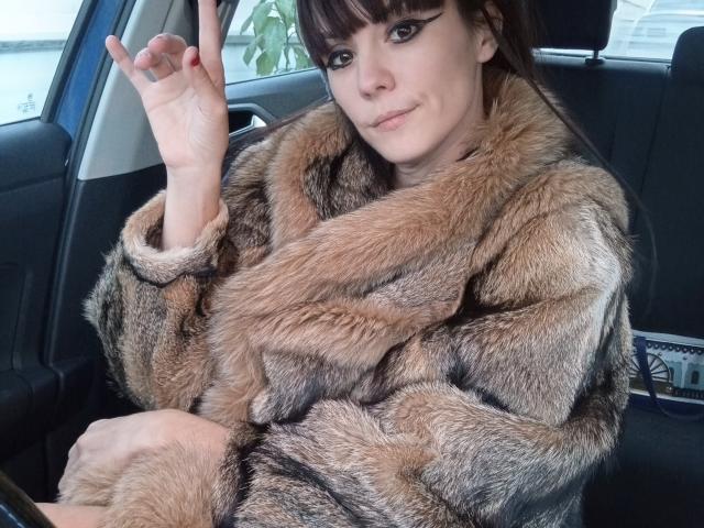 OrianaLaFrancaise, avec des cheveux bruns et une frange, portant un manteau en fourrure marron et noir, assise dans une voiture, faisant un signe de la paix, souriant légèrement..