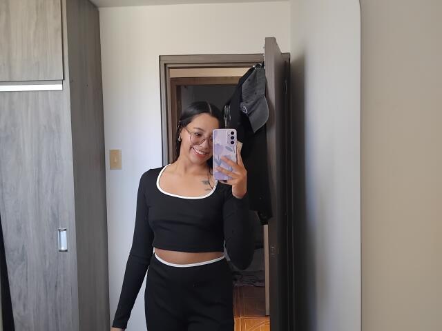 Mllevv with black crop top and matching pants, smiling, holding phone, showing a tattoo on her chest, standing in doorway, black jacket hanging.