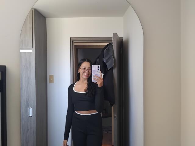 Mllevv has tits, wearing a black long-sleeve crop top and black pants, smiling in a mirror selfie with a phone, standing in a doorway with a hanging jacket.